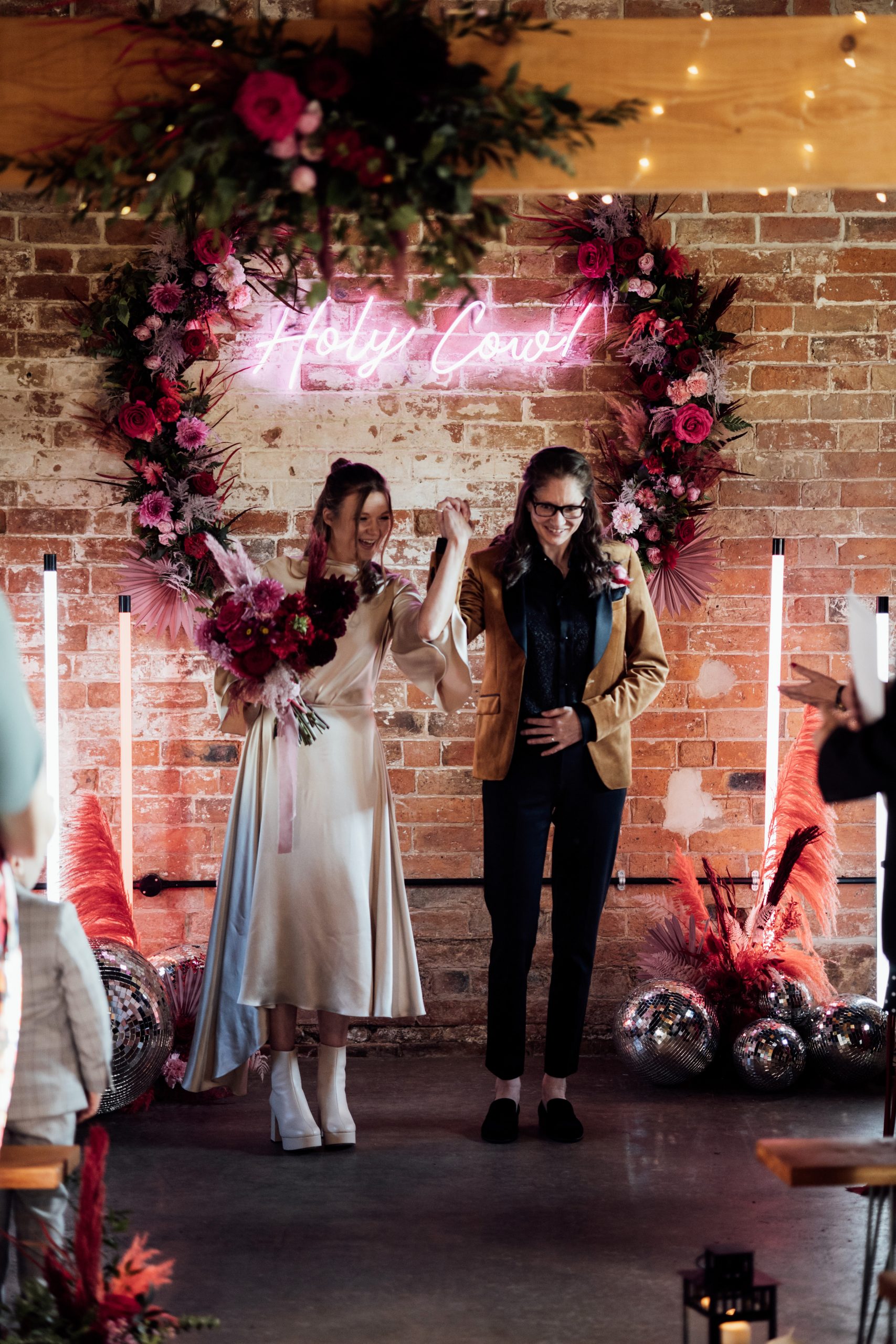 Two brides walking down the aisle with floral backdrop, disco balls and neon stands