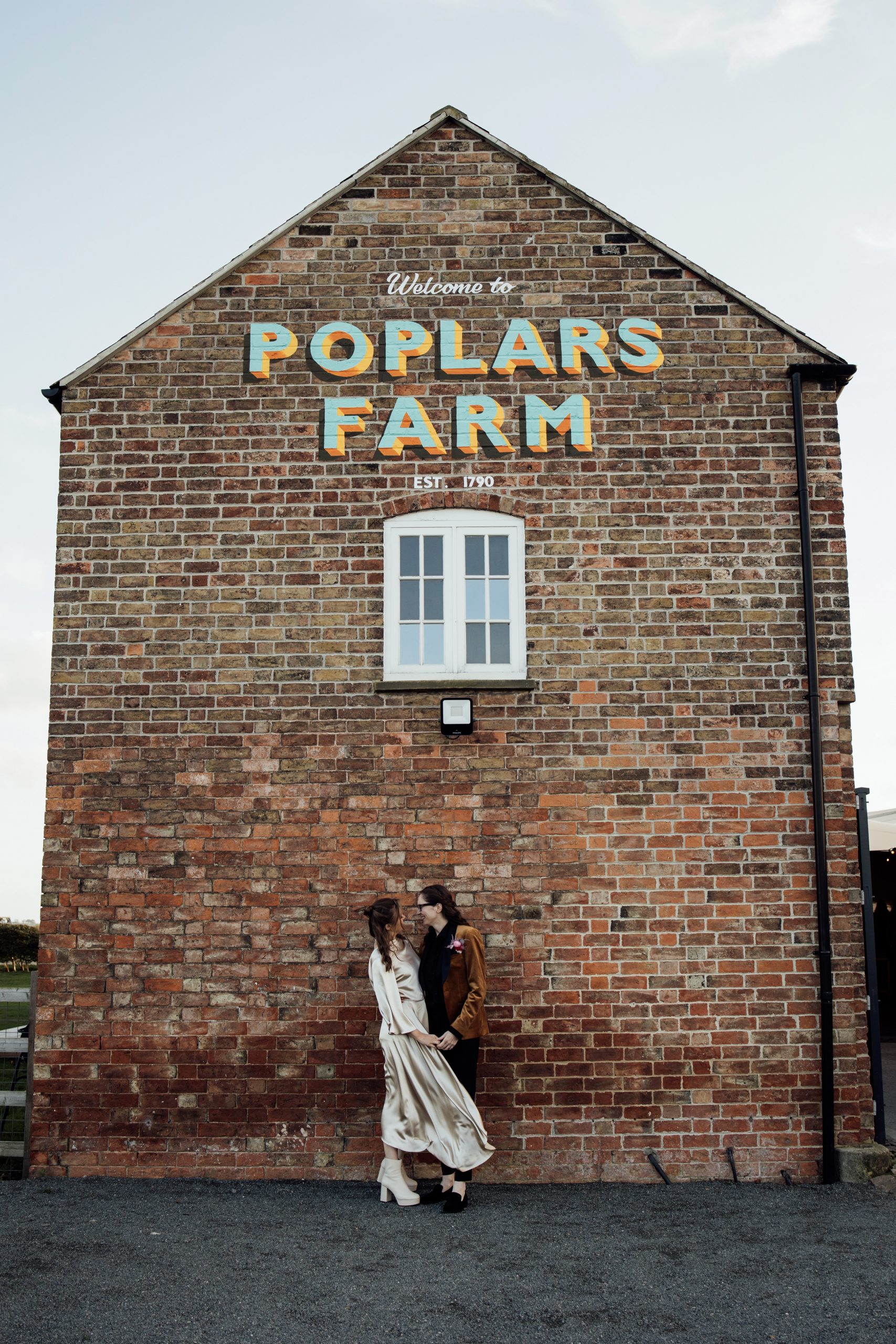 Two brides smiling under a vintage poplars farm sign outside a barn