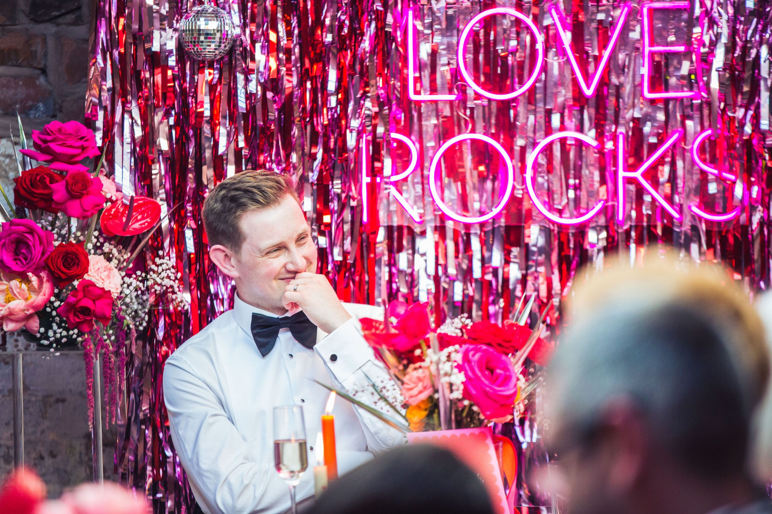 Groom sitting in front of a streamers backdrop with love rocks pink neon, flowers and disco balls
