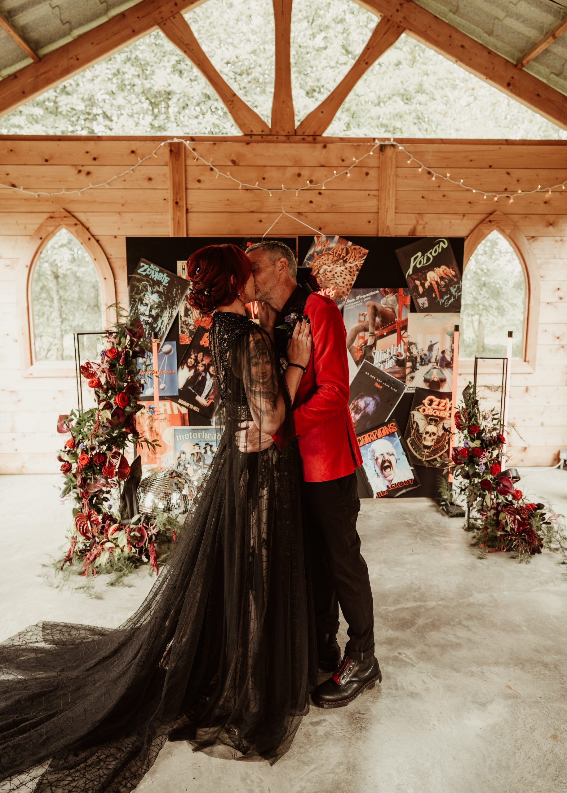 a bride in a black dress kissing her new husband who is wearing a red velvet jacket. Infront of a large poster board ceremony back drop.
