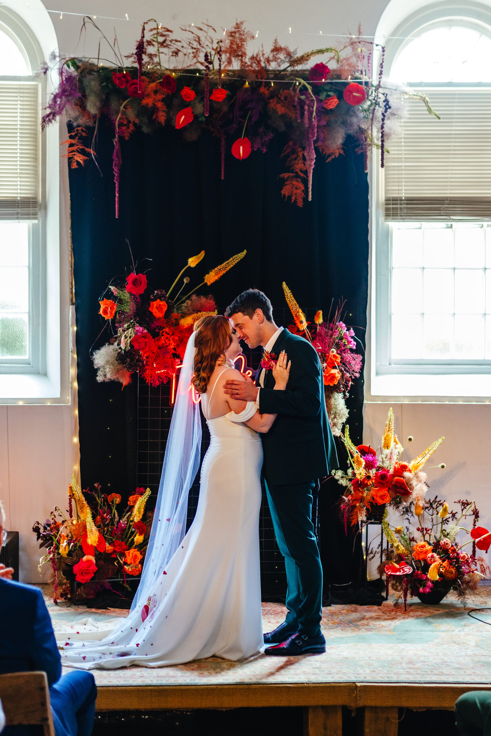 Bride and Groom kissing in front of black draping fabric and surrounded by colourful flowers with til death neon