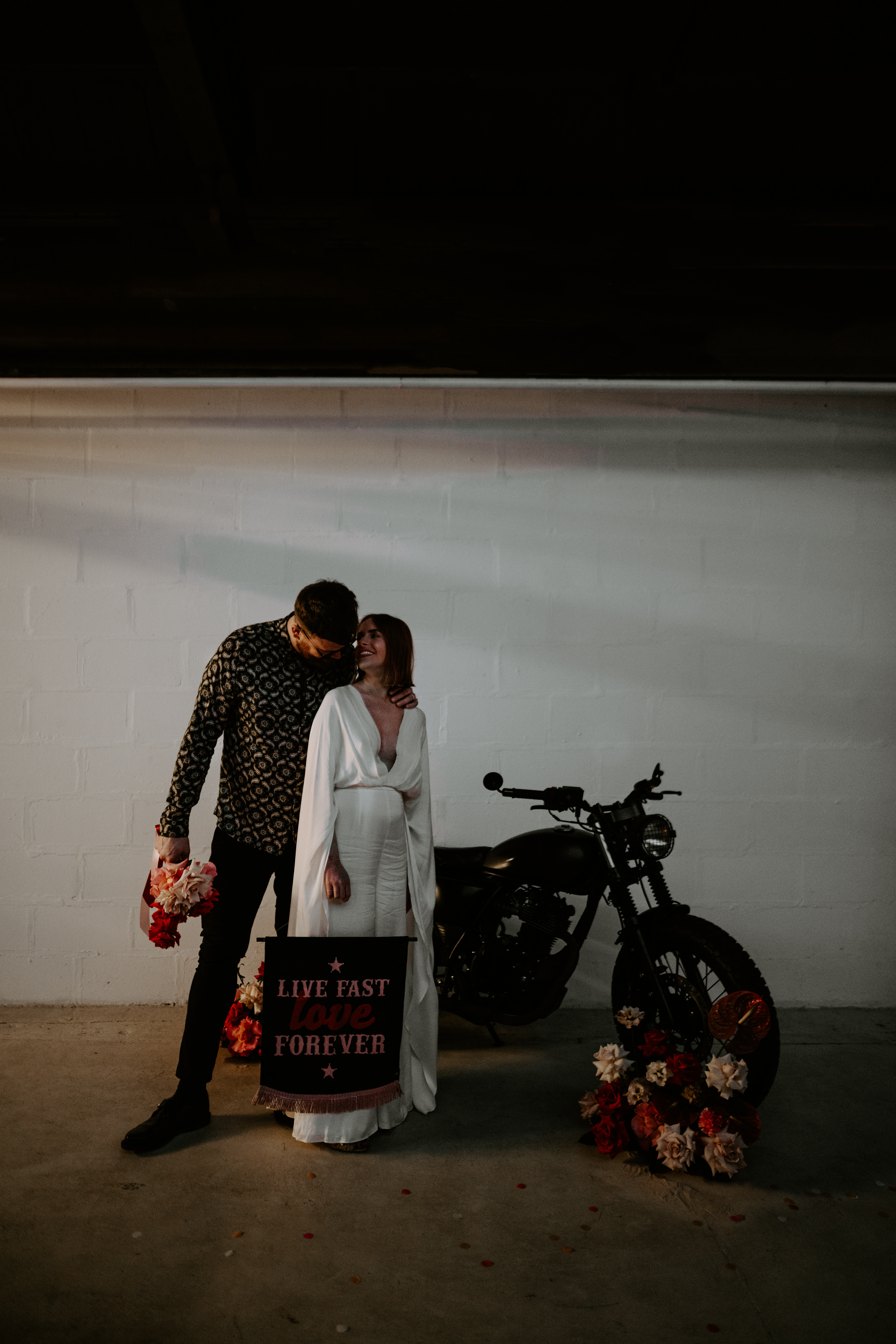 A bride and groom standing infront of a motorbike with pink and red flowers